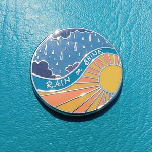 Puede incluir: Un pin de esmalte redondo con un cielo azul, nubes blancas y gotas de lluvia en el lado izquierdo. El lado derecho presenta un sol amarillo con rayos naranjas y el texto "RAIN or SHINE".