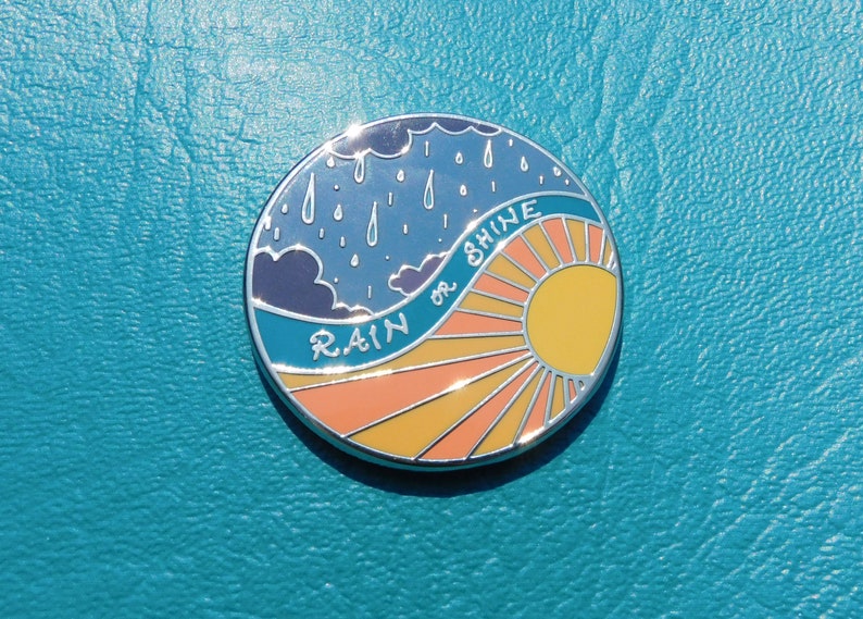 Puede incluir: Un pin de esmalte redondo con un cielo azul, nubes blancas y gotas de lluvia en el lado izquierdo. El lado derecho presenta un sol amarillo con rayos naranjas y el texto "RAIN or SHINE".