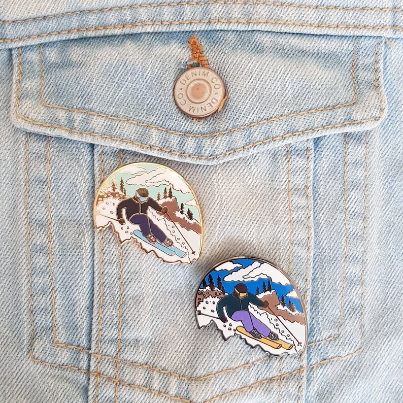 K&ouml;nnte beinhalten: Zwei Emaille-Pins mit Skifahrern auf einem schneebedeckten Berg. Die Pins sind golden und silbern mit blauen, wei&szlig;en und braunen Akzenten. Die Pins sind an einer blauen Jeansjacke befestigt.
