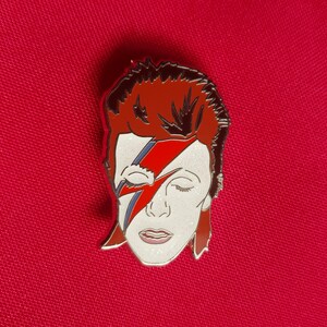 Puede incluir: Un pin de esmalte plateado con un retrato de David Bowie con su caracter&iacute;stico maquillaje de rayo rojo y azul. El pin es un primer plano de su rostro y cabello.