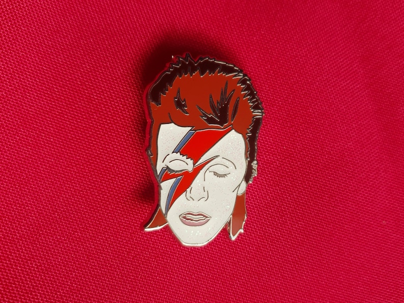 Puede incluir: Un pin de esmalte plateado con un retrato de David Bowie con su caracter&iacute;stico maquillaje de rayo rojo y azul. El pin es un primer plano de su rostro y cabello.