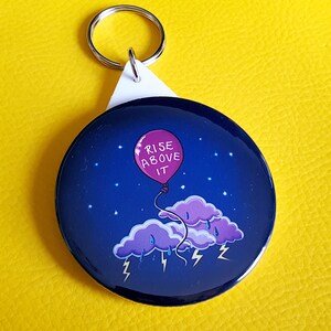 Puede incluir: Un llavero redondo con fondo azul y un globo rosa que dice "Rise Above It". El globo flota sobre una nube azul oscuro con rayos.