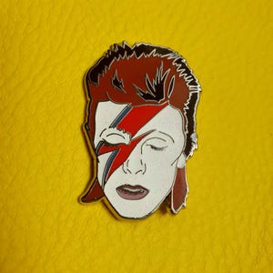 Puede incluir: Un pin de esmalte plateado con un retrato de David Bowie con su caracter&iacute;stico maquillaje de rayo. El pin tiene un dise&ntilde;o de rayo rojo, azul y blanco sobre un fondo blanco. El fondo del pin es rojo y negro.
