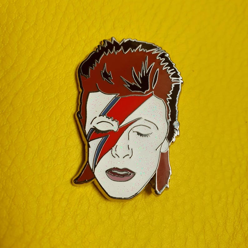 Puede incluir: Un pin de esmalte plateado con un retrato de David Bowie con su caracter&iacute;stico maquillaje de rayo. El pin tiene un dise&ntilde;o de rayo rojo, azul y blanco sobre un fondo blanco. El fondo del pin es rojo y negro.