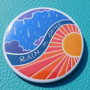 May include: A round button with a colourful design. One half of the button features a blue sky with white rain drops and purple clouds. The other half features a red and orange sun with rays. The text "RAIN OR SHINE" is written in a curved line on the button.