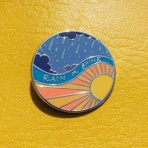Puede incluir: Un pin de esmalte redondo con un cielo azul con nubes de lluvia y un sol amarillo con rayos naranjas. El texto "RAIN or SHINE" est&aacute; escrito en una fuente blanca cursiva.