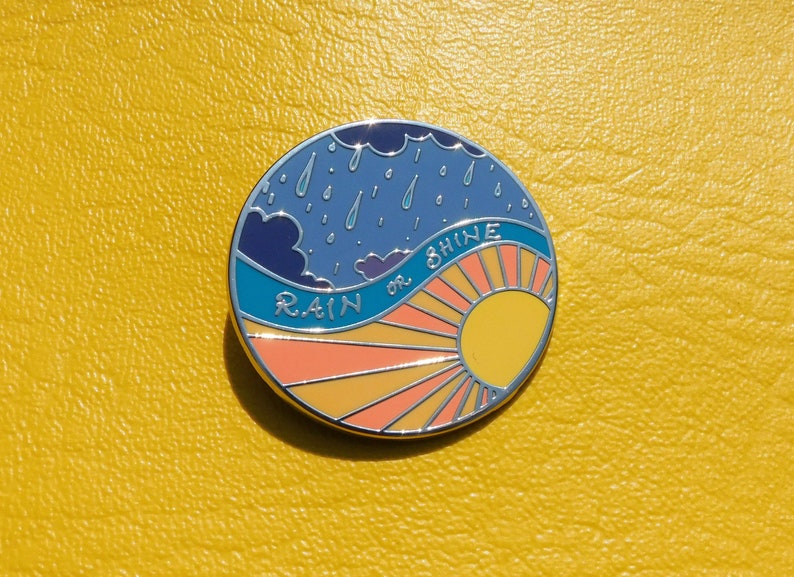 Puede incluir: Un pin de esmalte redondo con un cielo azul con nubes de lluvia y un sol amarillo con rayos naranjas. El texto "RAIN or SHINE" est&aacute; escrito en una fuente blanca cursiva.