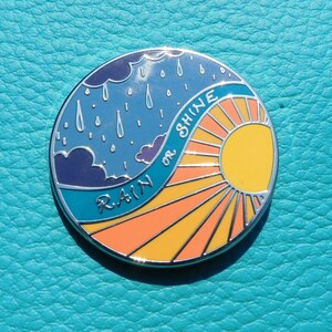 Puede incluir: Un pin redondo de metal con un cielo azul y nubes de lluvia en un lado y un sol amarillo con rayos naranjas en el otro. El texto "RAIN or SHINE" est&aacute; escrito en una l&iacute;nea curva en el pin.