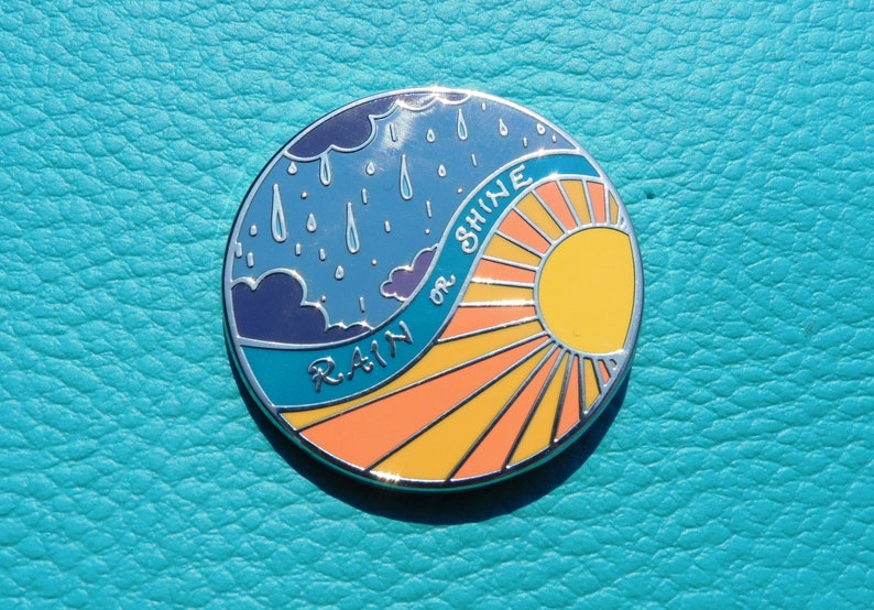 Puede incluir: Un pin redondo de metal con un cielo azul y nubes de lluvia en un lado y un sol amarillo con rayos naranjas en el otro. El texto "RAIN or SHINE" est&aacute; escrito en una l&iacute;nea curva en el pin.