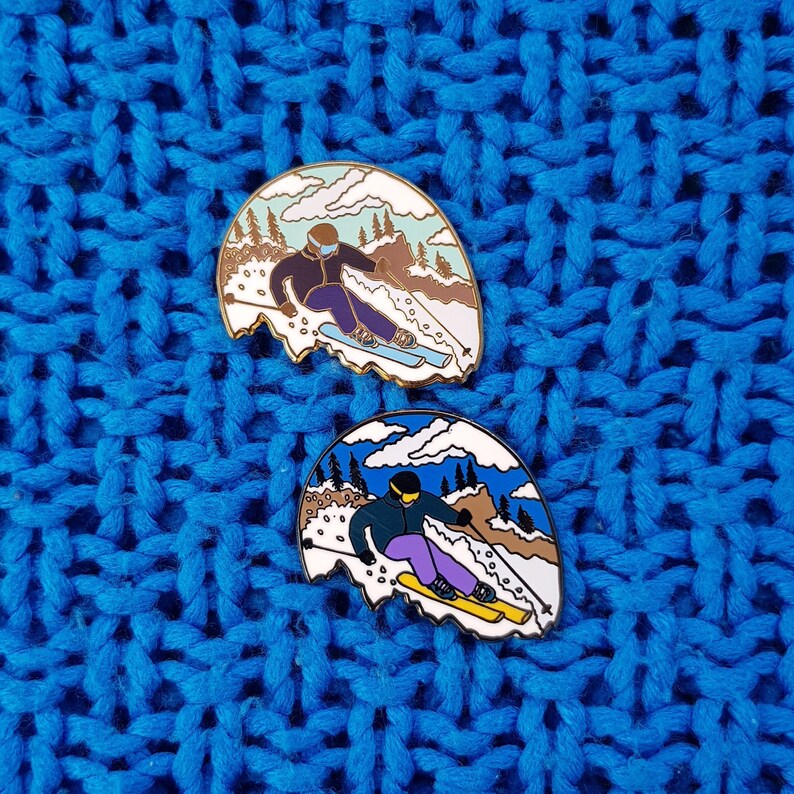 K&ouml;nnte beinhalten: Zwei Emaille-Pins mit Skifahrern vor einem Bergpanorama. Die Pins sind wei&szlig; und blau mit goldenen Akzenten. Die Skifahrer tragen blaue und wei&szlig;e Skianz&uuml;ge und halten Skist&ouml;cke.