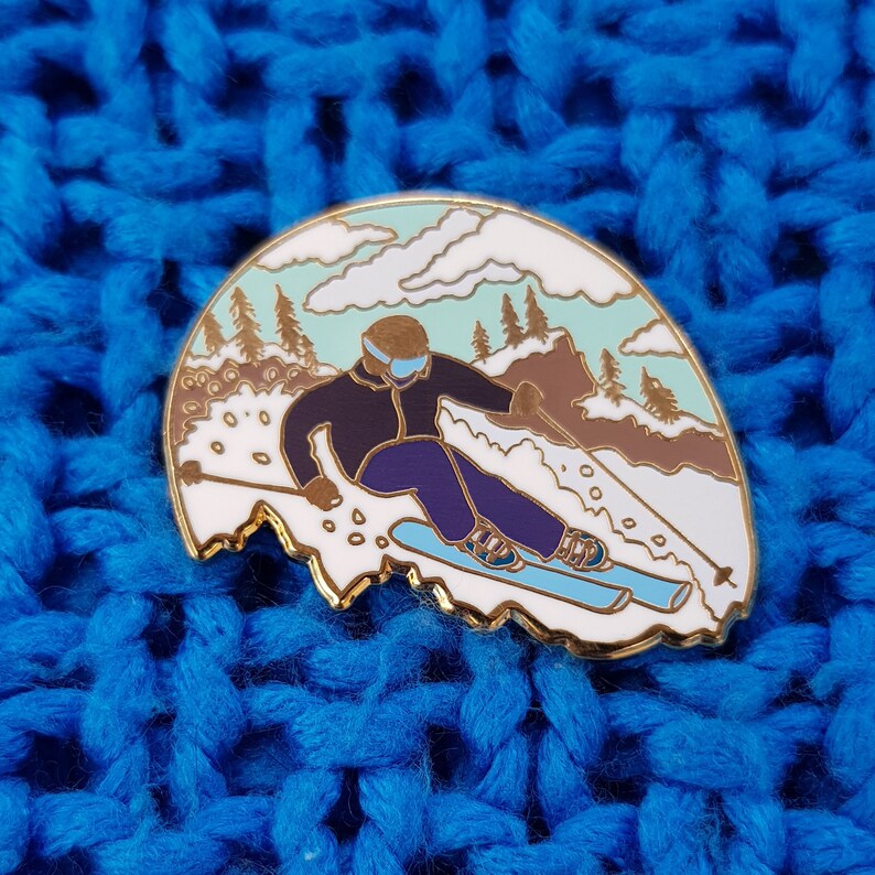 K&ouml;nnte beinhalten: Eine goldfarbene Emaille-Anstecknadel mit einem Skifahrer in einer schwarzen Jacke und lila Hose, der einen schneebedeckten Berg mit blauem Himmel und wei&szlig;en Wolken im Hintergrund hinunterf&auml;hrt.
