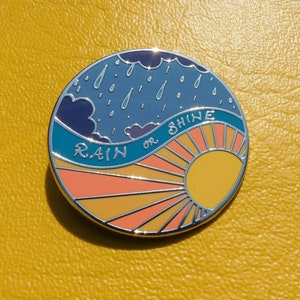 Puede incluir: Un pin de esmalte redondo con un cielo azul, nubes blancas y gotas de lluvia en el lado izquierdo. El lado derecho presenta un sol amarillo con rayos naranjas y el texto "RAIN OR SHINE".