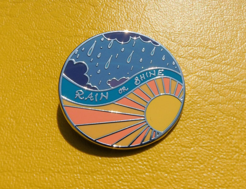 Puede incluir: Un pin de esmalte redondo con un cielo azul, nubes blancas y gotas de lluvia en el lado izquierdo. El lado derecho presenta un sol amarillo con rayos naranjas y el texto "RAIN OR SHINE".