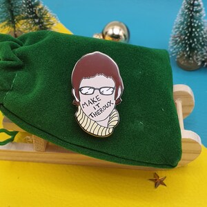 May include: A green velvet pouch with a gold pin featuring a cartoon illustration of a person with brown hair wearing glasses and a yellow scarf. The text "MAKE IT THERO" is written on the scarf.