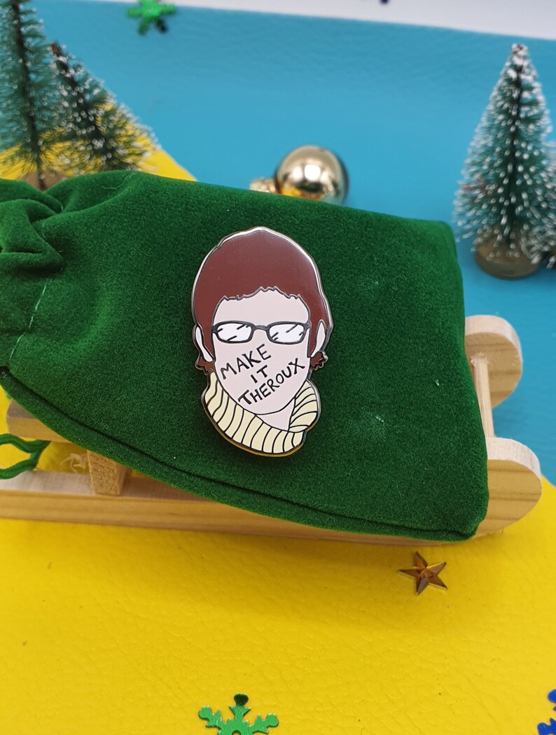 May include: A green velvet pouch with a gold pin featuring a cartoon illustration of a person with brown hair wearing glasses and a yellow scarf. The text "MAKE IT THERO" is written on the scarf.