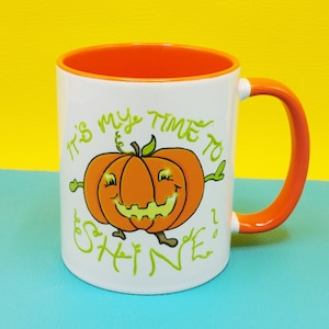 May include: White ceramic mug with an orange rim and handle. The mug features a cartoon pumpkin with a smiling face and the text "It's my time to shine!"