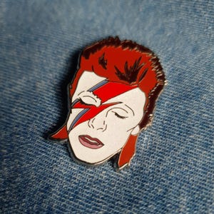 Puede incluir: Un pin de esmalte plateado con una ilustraci&oacute;n roja y azul de la cara de David Bowie con su caracter&iacute;stico maquillaje de rayo.