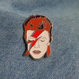 Puede incluir: Un pin de esmalte que representa un retrato de David Bowie con su ic&oacute;nico maquillaje de rayo, en rojo y blanco.