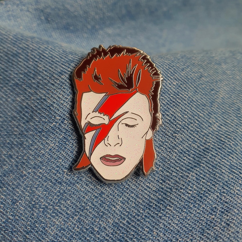 Puede incluir: Un pin de esmalte que representa un retrato de David Bowie con su ic&oacute;nico maquillaje de rayo, en rojo y blanco.