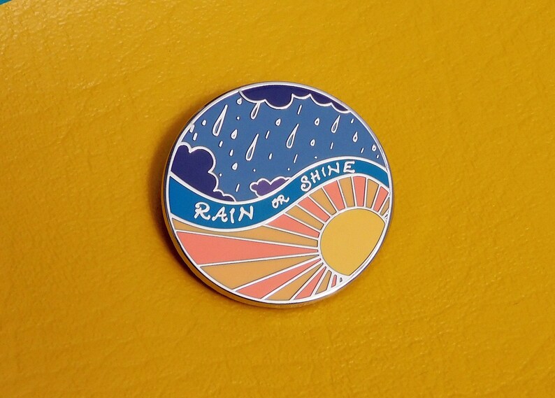 Puede incluir: Un pin de esmalte redondo con un cielo azul, nubes blancas y gotas de lluvia en la mitad superior. La mitad inferior es una salida del sol con rayos naranja, amarillo y rosa. El texto "RAIN or SHINE" est&aacute; escrito en blanco sobre una ola azul.