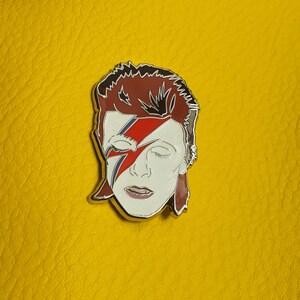 Puede incluir: Un pin de esmalte plateado con un retrato de David Bowie con su caracter&iacute;stico maquillaje de rayo. El pin est&aacute; sobre un fondo amarillo.