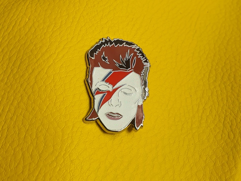 Puede incluir: Un pin de esmalte plateado con un retrato de David Bowie con su caracter&iacute;stico maquillaje de rayo. El pin est&aacute; sobre un fondo amarillo.