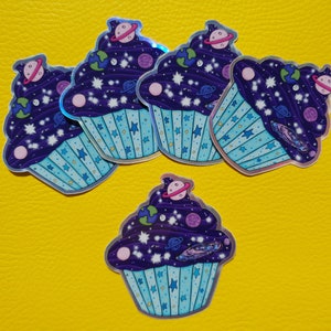 May include: Five holographic cupcake stickers with a galaxy theme. The cupcakes are purple with blue frosting and white stars. Each cupcake has a different planet or celestial body on top.