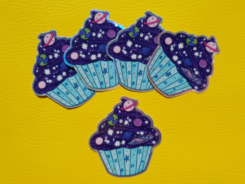May include: Five holographic cupcake stickers with a galaxy theme. The cupcakes are purple with blue frosting and white stars. Each cupcake has a different planet or celestial body on top.