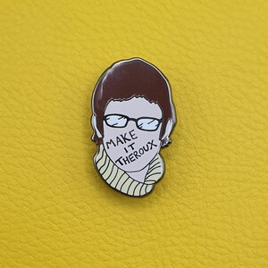 May include: A brown and white enamel pin featuring a cartoon illustration of a person with brown hair wearing glasses and a yellow and white striped sweater. The person's mouth is covered with the text "MAKE IT THEROUX".