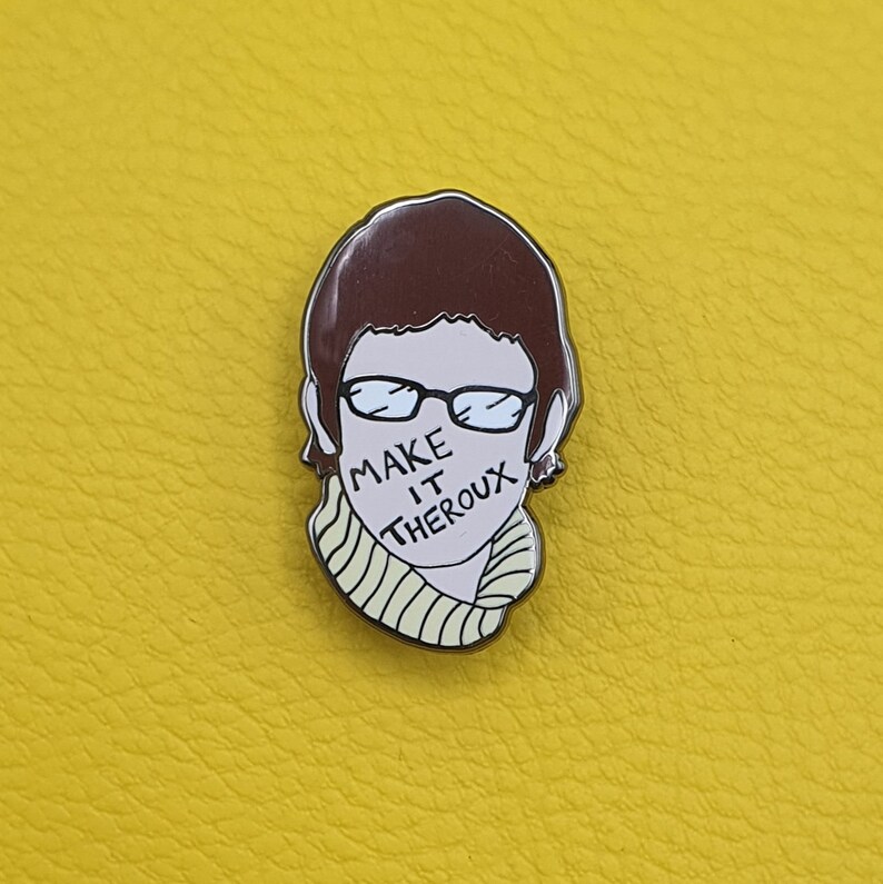 May include: A brown and white enamel pin featuring a cartoon illustration of a person with brown hair wearing glasses and a yellow and white striped sweater. The person's mouth is covered with the text "MAKE IT THEROUX".