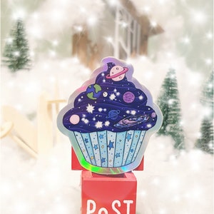 May include: A holographic sticker of a blue cupcake with a galaxy design and planets. The sticker is on a red mailbox with the word "POST" written on it.
