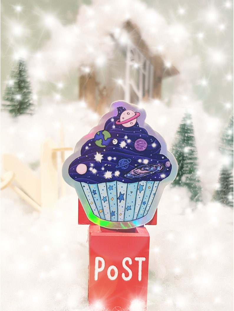 May include: A holographic sticker of a blue cupcake with a galaxy design and planets. The sticker is on a red mailbox with the word "POST" written on it.