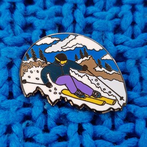 K&ouml;nnte beinhalten: Eine farbige Emaille-Anstecknadel, die einen Skifahrer in einer blauen Jacke und einer lila Hose zeigt, der einen schneebedeckten Berg mit blauem Himmel und wei&szlig;en Wolken im Hintergrund hinunterf&auml;hrt.