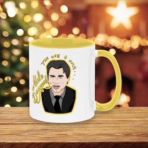 May include: White ceramic mug with a yellow rim and handle. The mug features a cartoon illustration of a man with the text "The one & only...  Adele Dazeem!"
