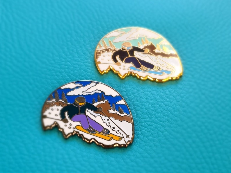 K&ouml;nnte beinhalten: Zwei goldfarbene Emaille-Pins, die einen Skifahrer in einer Winterlandschaft zeigen. Die Pins sind in Form eines Gebirges mit blauem Himmel und wei&szlig;en Wolken gestaltet. Der Skifahrer tr&auml;gt eine schwarze Jacke, eine lila Hose und einen blauen Helm. Der Skifahrer h&auml;lt Skist&ouml;cke und f&auml;hrt einen Hang hinunter. Die Pins sind eine lustige und einzigartige M&ouml;glichkeit, Ihre Liebe zum Wintersport zu zeigen.