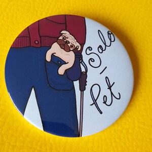 May include: A white button featuring a cartoon illustration of a person wearing a red shirt and blue trousers with a small brown and white dog peeking out of the pocket. The text "Pet-Salo" is written in black cursive on the button.
