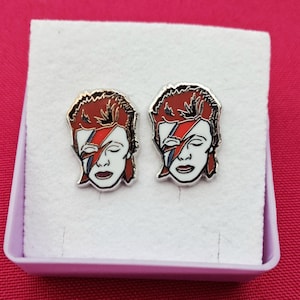 Puede incluir: Un par de pendientes de botón plateados con un retrato estilizado de una persona con cabello rojo y un rayo en la cara. Los pendientes se presentan en una caja blanca con interior rosa, sobre un fondo rosa.