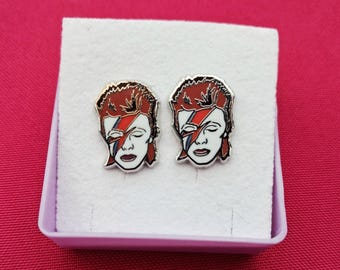 May include: A pair of silver-toned stud earrings featuring a stylized portrait of a person with red hair and a lightning bolt across the face. The earrings are presented in a white box with a pink interior, set against a pink background.