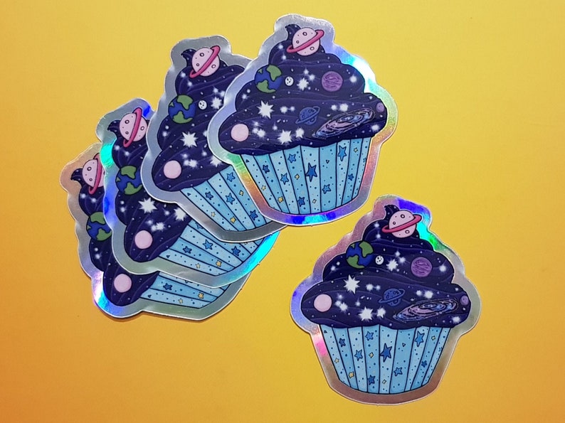 May include: Four iridescent stickers of a cupcake with a blue frosting and a galaxy design. The cupcake is filled with stars, planets, and a galaxy swirl.