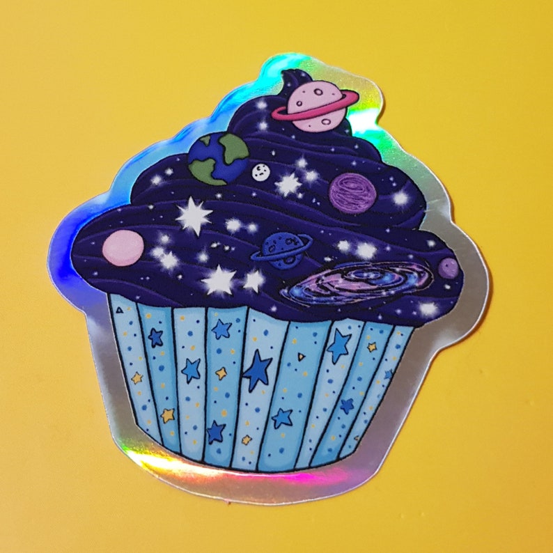 May include: A holographic sticker of a cupcake with a blue and purple galaxy frosting and a blue and white striped base. The frosting is decorated with stars, planets, and a galaxy swirl.
