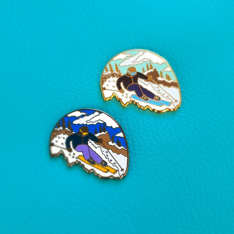 K&ouml;nnte beinhalten: Zwei Emaille-Pins, die Skifahrer in einer Winterlandschaft zeigen. Die Pins sind goldfarben mit wei&szlig;en, blauen und braunen Akzenten. Die Skifahrer tragen blaue und lilafarbene Skianz&uuml;ge und fahren einen schneebedeckten Hang hinunter.
