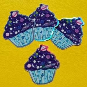 May include: Four holographic stickers of galaxy cupcakes. Each cupcake is decorated with a purple galaxy frosting with white stars, planets, and a galaxy swirl. The cupcakes are on a yellow background.