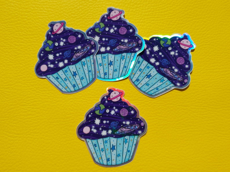 May include: Four holographic stickers of galaxy cupcakes. Each cupcake is decorated with a purple galaxy frosting with white stars, planets, and a galaxy swirl. The cupcakes are on a yellow background.
