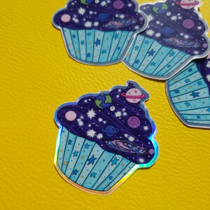 May include: A holographic sticker of a blue cupcake with a purple frosting and a galaxy design. The frosting is filled with stars, planets, and a galaxy swirl.