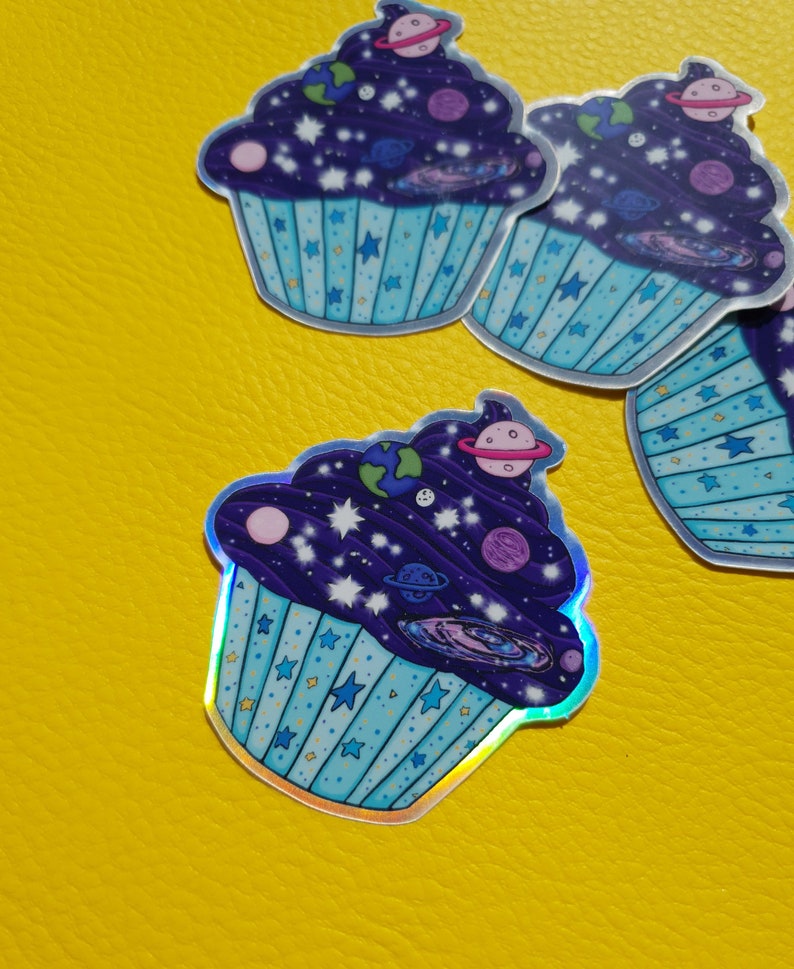 May include: A holographic sticker of a blue cupcake with a purple frosting and a galaxy design. The frosting is filled with stars, planets, and a galaxy swirl.