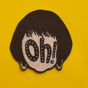 Op de afbeelding: Gestikt patch van een cartoonfiguur met bruin haar en een roze gezicht. Het personage zegt "Oh!" in zwarte letters met witte stippen.