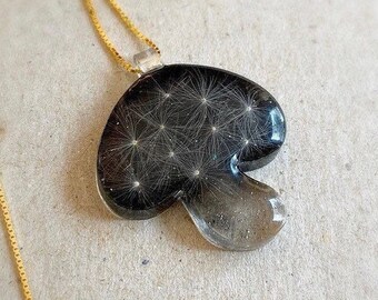 May include: A unique pendant necklace featuring a mushroom-shaped charm. The charm is black with embedded white dandelion seeds, suspended in clear resin. The necklace has a delicate gold chain.