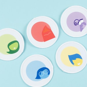 May include: Five white, circular coasters with cartoon character designs in various colours. Each coaster represents a different emotion: green, red, purple, yellow, and blue. The coasters are set against a light blue background.