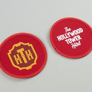 Op de afbeelding: Twee rode ronde patches. Eén heeft een gouden embleem met de letters "HTH" in een schild. De andere patch heeft de tekst "The Hollywood Tower Hotel" in het wit. Beide patches hebben een rode rand.
