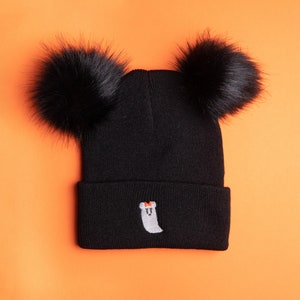 May include: Black knitted beanie hat with two large black faux fur pom-poms. The hat has an embroidered ghost with a small orange bow. The beanie is set against an orange background, creating a Halloween theme.